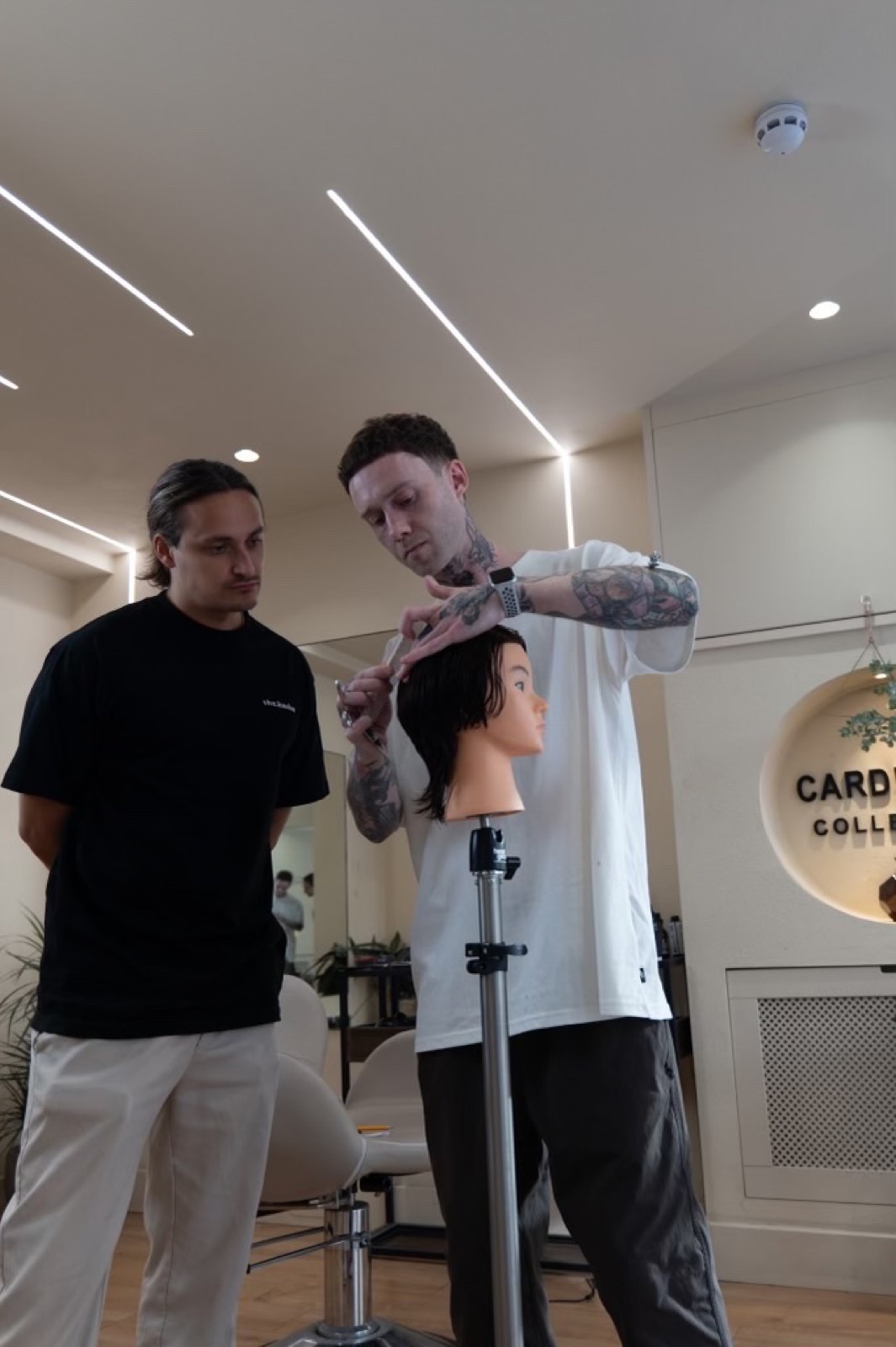 A stylist cutting a training head while a student watches closely, inside the Cardinale Collective salon